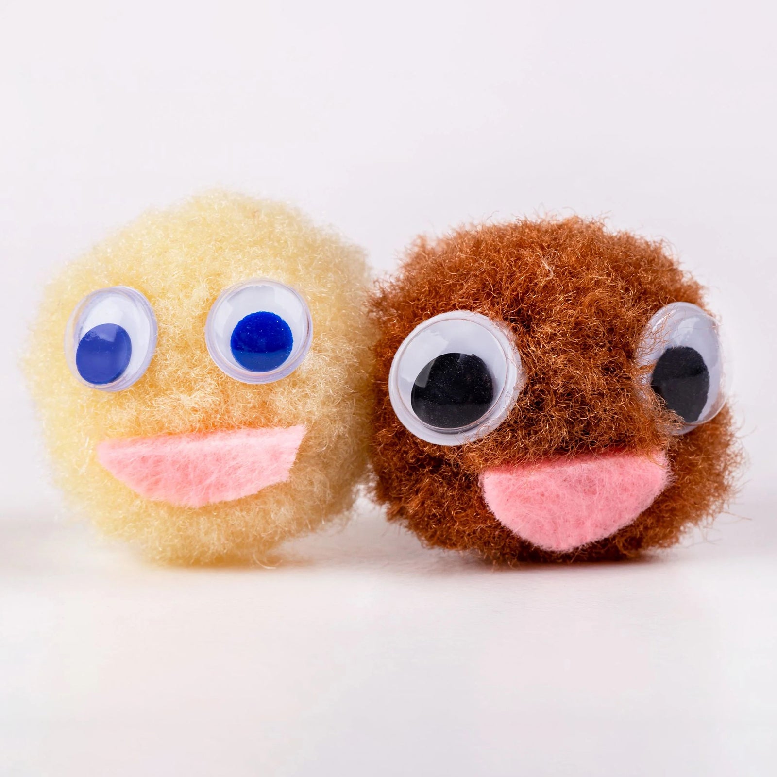Close up image of two pom pom faces created with different skin tone colours and large joggle eyes. The image highlights the texture, softness and creative potential of the pom poms for character making, storytelling activities and diversity focused craft projects.