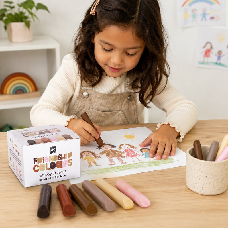 A young child uses a brown Friendship Colours Stubby Crayon to draw a family picture at a table. The image demonstrates how the chunky crayons fit comfortably in small hands while encouraging creative expression and inclusive artwork.
