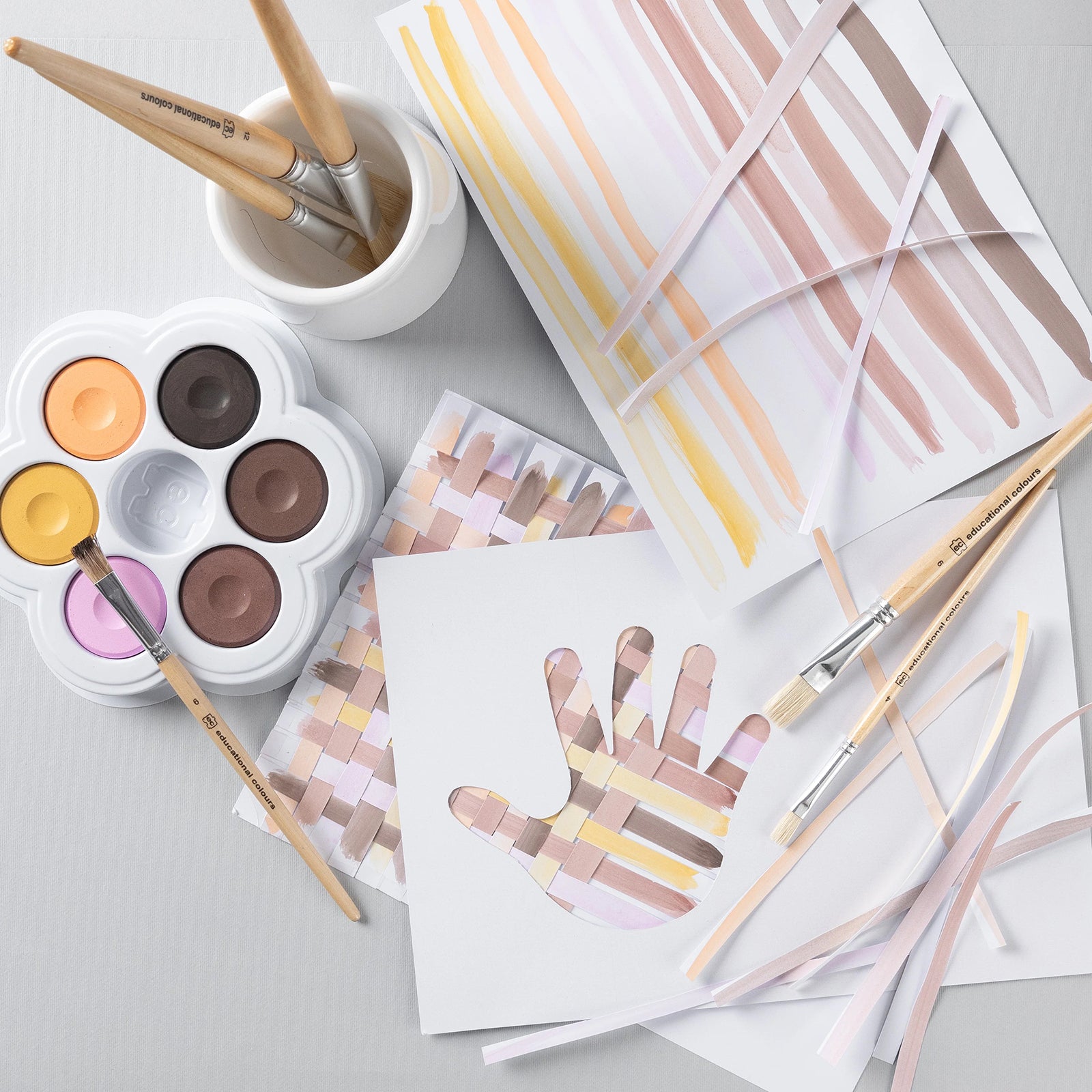The palette is featured within a creative craft activity involving painted paper strips and weaving projects. Brushes, painted samples and craft materials demonstrate the versatility of the watercolours across multiple educational art applications.