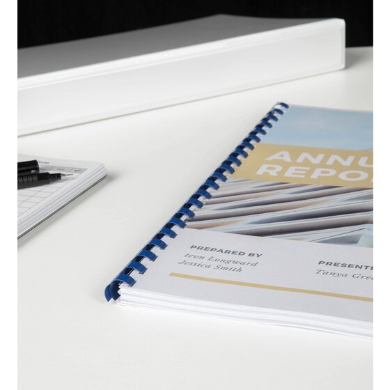 Annual report bound with a blue plastic comb on a white desk, with a notepad and pens beside it