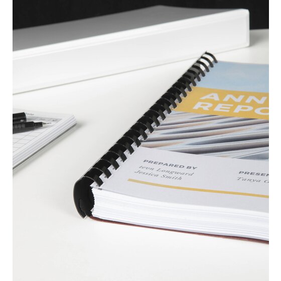 Annual Report bound along the left edge with a black GBC plastic comb, lying on a white desk beside a white binder and pens