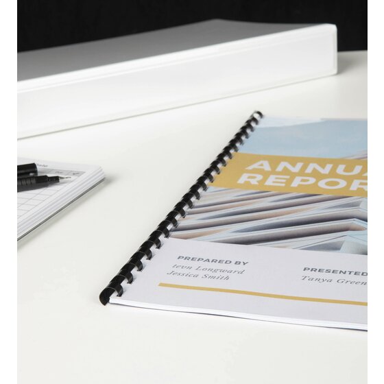 Annual report bound with a black plastic comb on a white desk, with a white ring binder and pens in the background