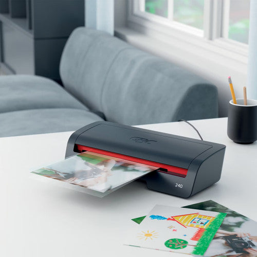 Black and red GBC 240 laminator on a white desk by a window laminating a photo, with a child drawing beside it