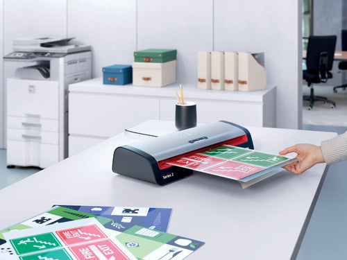Hand feeding a laminated safety sign into a GBC 250HS on a white office desk, with a copier, shelving and more signs in the background