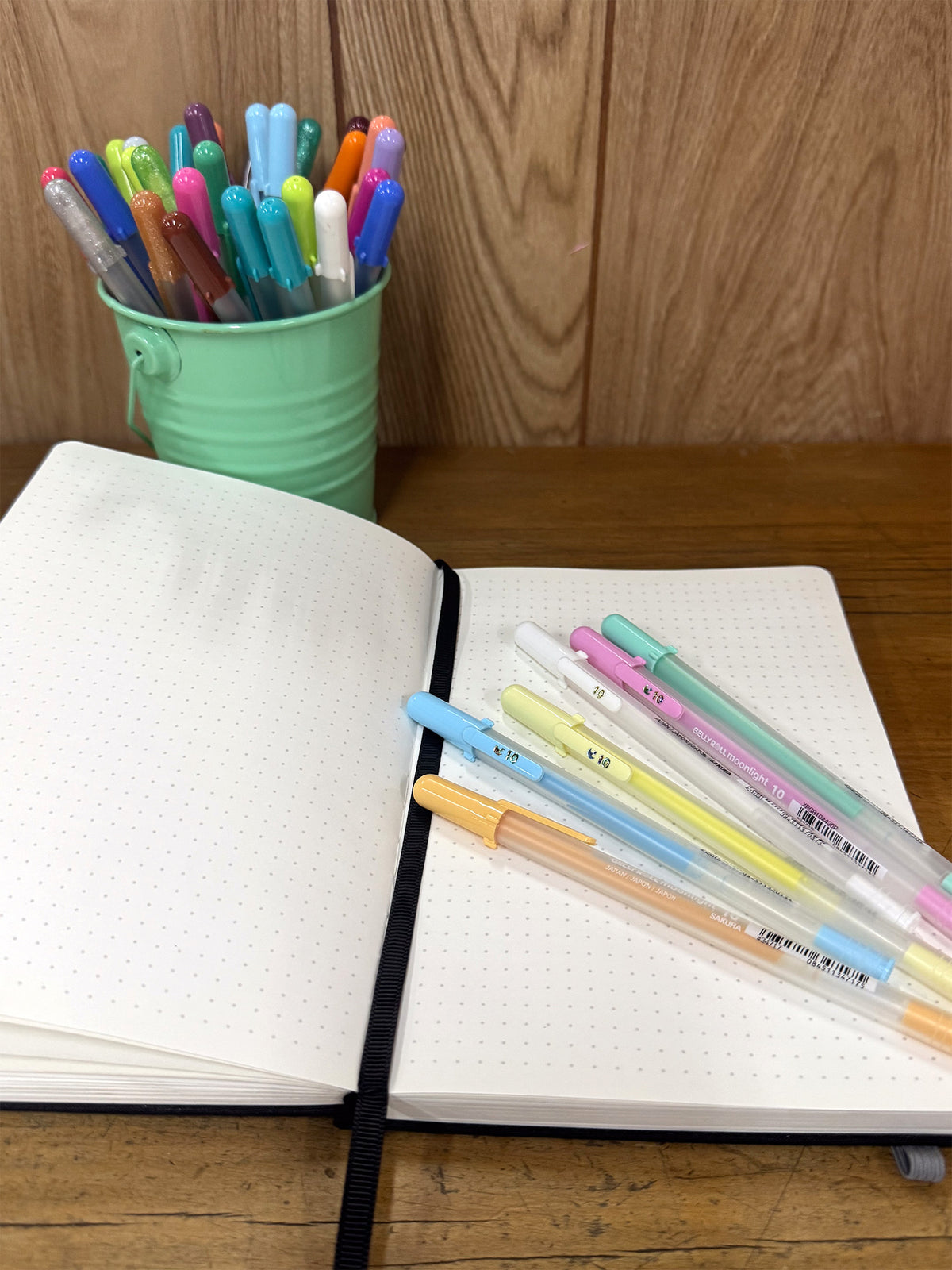 Open dotted notebook with pastel Moonlight pens and a mint pen pot on a wooden desk