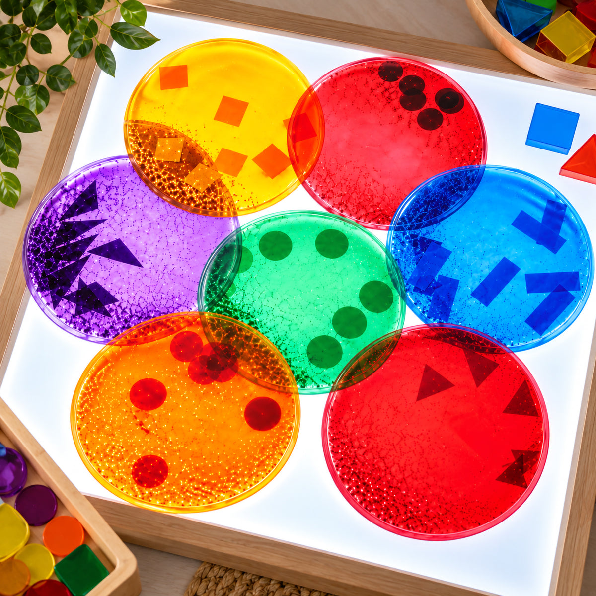Overhead view of seven colourful Giant Squidgy Sparkle Circles arranged across an illuminated light panel. The translucent sensory circles overlap to reveal colour interactions while floating circles, squares, triangles and rectangular shapes provide opportunities for visual observation, matching and early shape recognition.