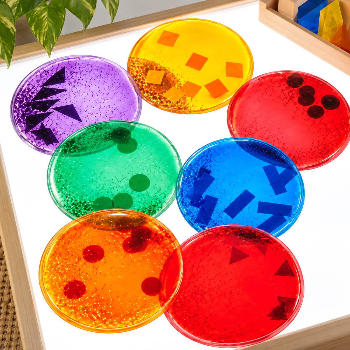 Close overhead classroom view of the Giant Squidgy Sparkle Circles glowing on a bright light panel. Seven assorted translucent circles display sparkling liquid and contrasting floating geometric shapes, creating an engaging sensory resource for exploring colour, transparency, patterns and visual effects.