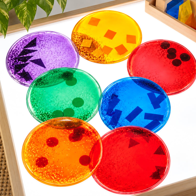 Close overhead classroom view of the Giant Squidgy Sparkle Circles glowing on a bright light panel. Seven assorted translucent circles display sparkling liquid and contrasting floating geometric shapes, creating an engaging sensory resource for exploring colour, transparency, patterns and visual effects.