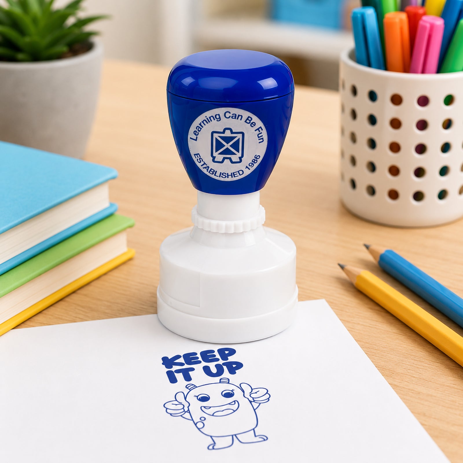 Close up view of the blue self inking merit stamp with books, pencils and classroom resources in the background. The Keep It Up impression demonstrates the large, easy to read design suitable for worksheets, assignments and student workbooks.