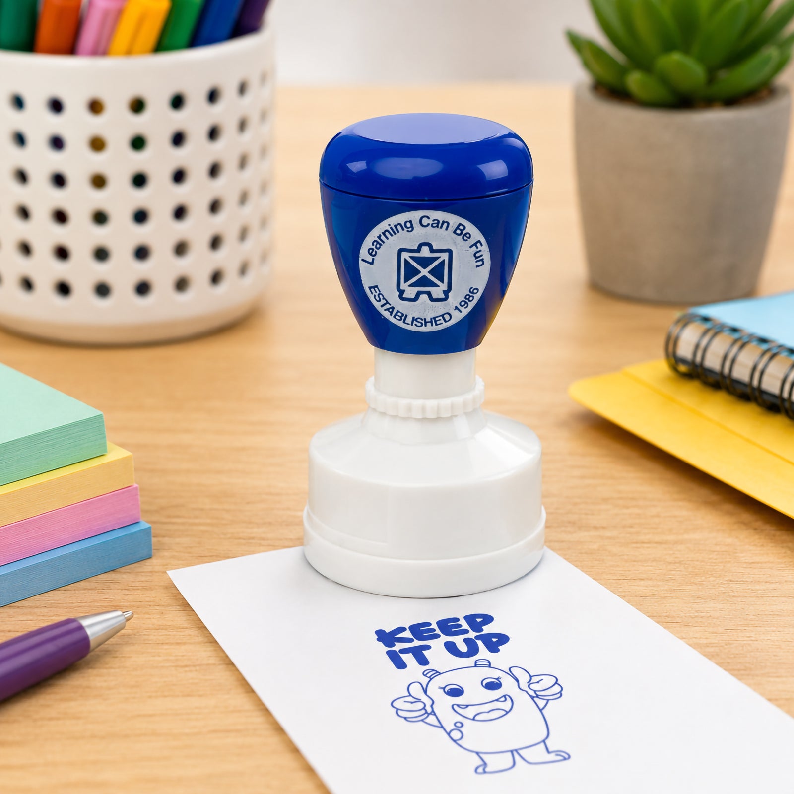 Learning Can Be Fun Keep It Up reward stamp positioned on a classroom desk surrounded by colourful notes, pens and learning materials. The bold blue stamp impression highlights the motivational message and friendly character designed to inspire confidence and persistence.