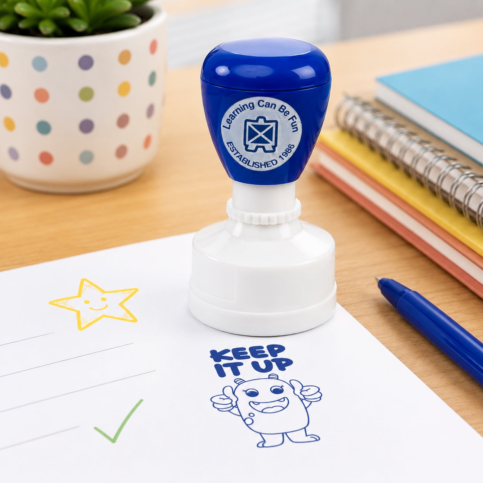Blue self inking Keep It Up merit stamp displayed on a student worksheet beside stationery and classroom supplies. The clear blue impression features a cheerful character giving two thumbs up, making it ideal for encouraging students and recognising ongoing effort.