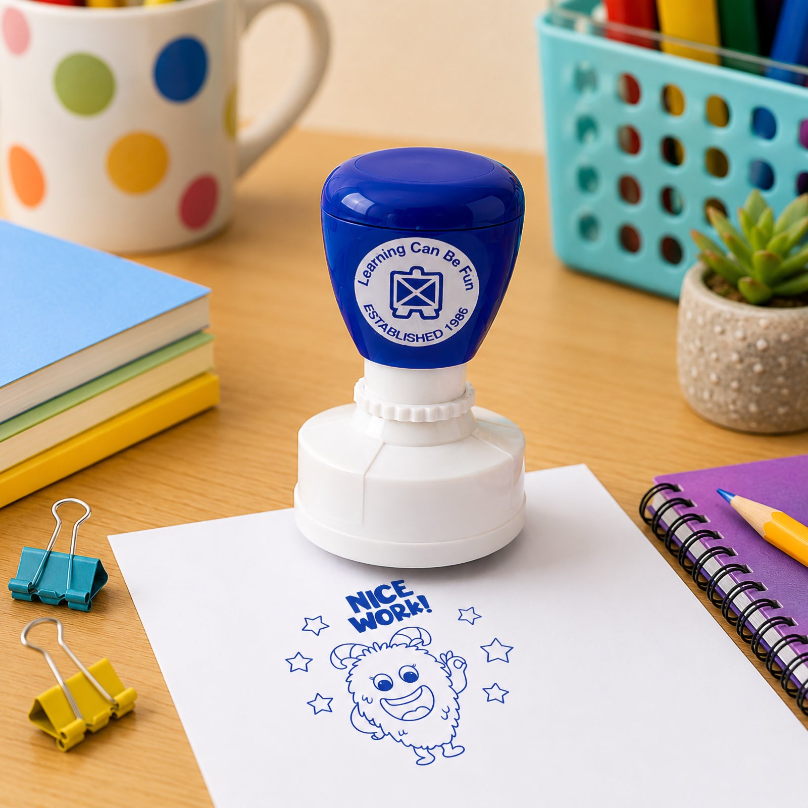 Close up classroom scene featuring the self inking Nice Work merit stamp with notebooks, pencil holders and learning supplies. The large blue impression demonstrates clear visibility and suitability for marking homework, worksheets and classroom activities.