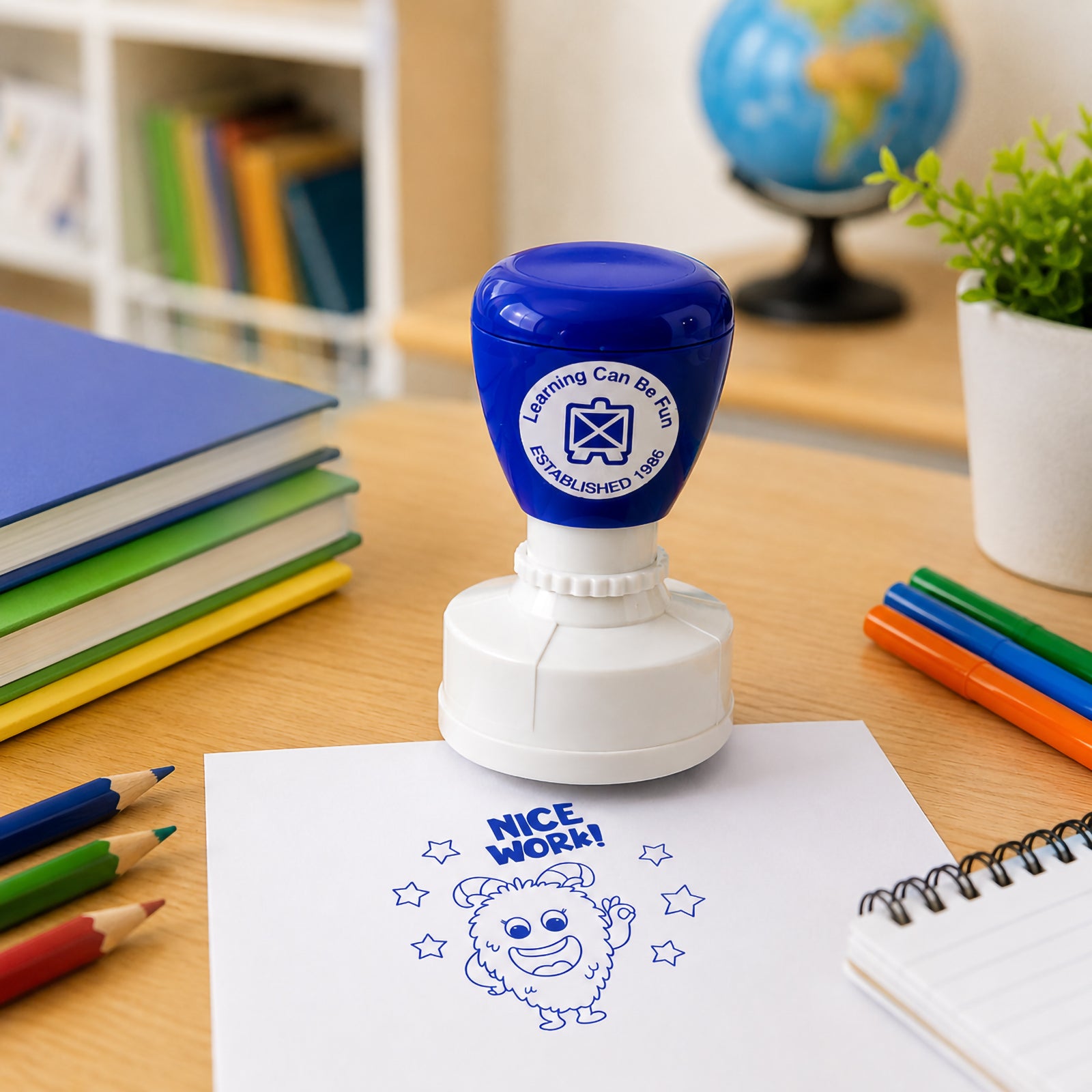 Blue self inking Nice Work merit stamp positioned on a classroom desk with books, pencils and educational resources. The stamped impression features a cheerful furry character surrounded by stars, making it ideal for rewarding student achievement and recognising quality work.