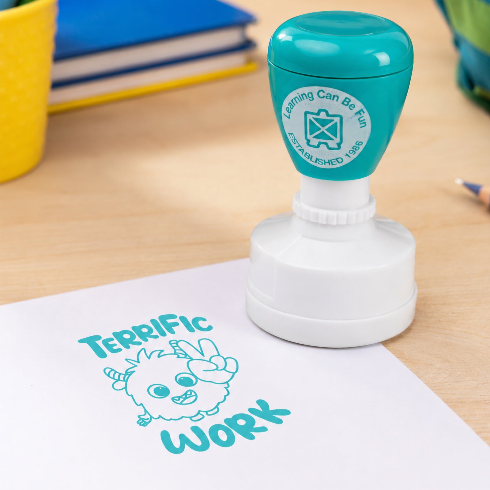 Close up classroom scene featuring the self inking merit stamp with books, pencil cases and educational resources in the background. The bold Terrific Work impression demonstrates the large and highly visible design suitable for worksheets, homework and classroom activities.