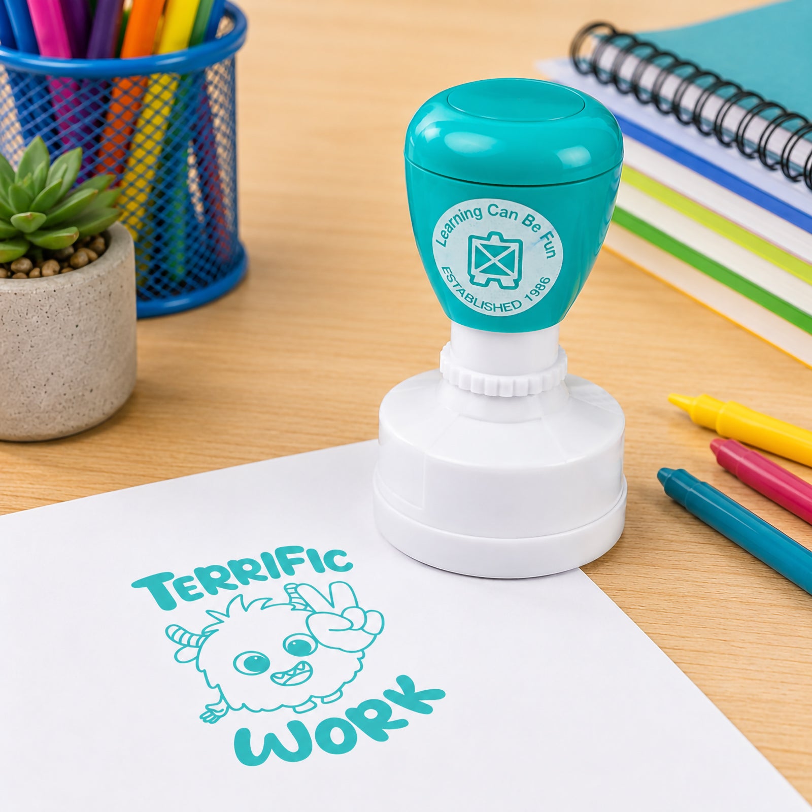 Teal self inking Terrific Work merit stamp displayed on a classroom desk beside colourful markers, notebooks and stationery. The clear stamped impression features a cheerful fuzzy character making a victory sign, helping teachers recognise excellent student achievement and effort.