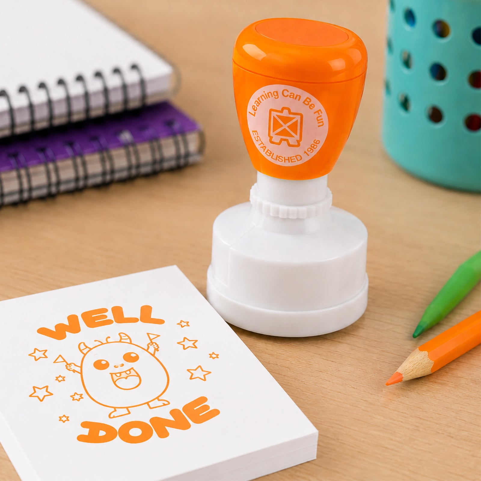 Close up classroom scene featuring the self inking Well Done merit stamp alongside notebooks, pencils and colourful learning supplies. The large orange impression demonstrates clear visibility and suitability for marking homework, assignments and student work.