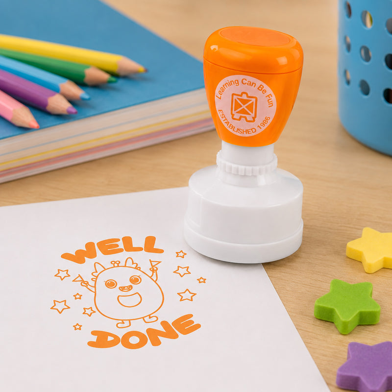 Orange self inking Well Done merit stamp displayed on a classroom desk beside coloured pencils, notebooks and star shaped erasers. The bright orange impression features a cheerful character celebrating among stars, making it ideal for recognising student achievement and progress.