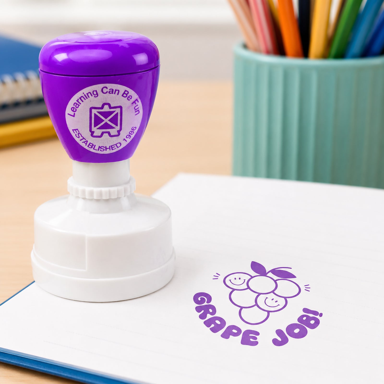 Close up view of the Grape Job reward stamp on an open workbook. The vibrant purple grape illustration stands out clearly, making positive feedback engaging and memorable for students.