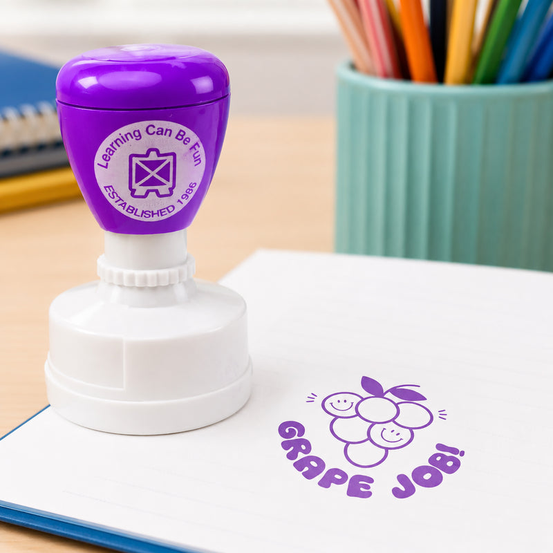 Close up view of the Grape Job reward stamp on an open workbook. The vibrant purple grape illustration stands out clearly, making positive feedback engaging and memorable for students.