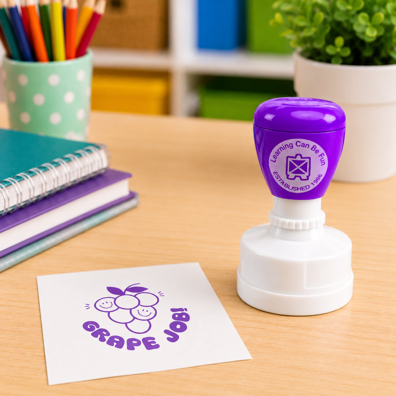 Purple self inking Grape Job merit stamp positioned beside notebooks and classroom supplies. The cheerful grape character design and bold purple impression help recognise effort, participation and learning success.