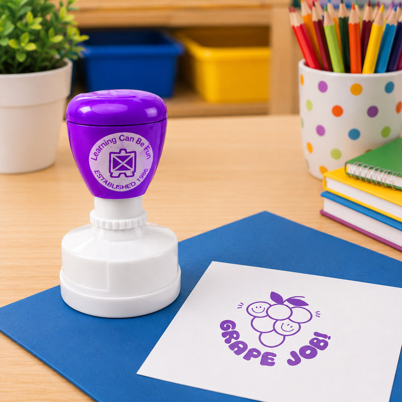 Learning Can Be Fun Grape Job Merit Stamp displayed on a classroom desk with colourful pencils and exercise books. The purple grape themed impression appears clearly on white paper, creating a fun and encouraging reward for student achievement.