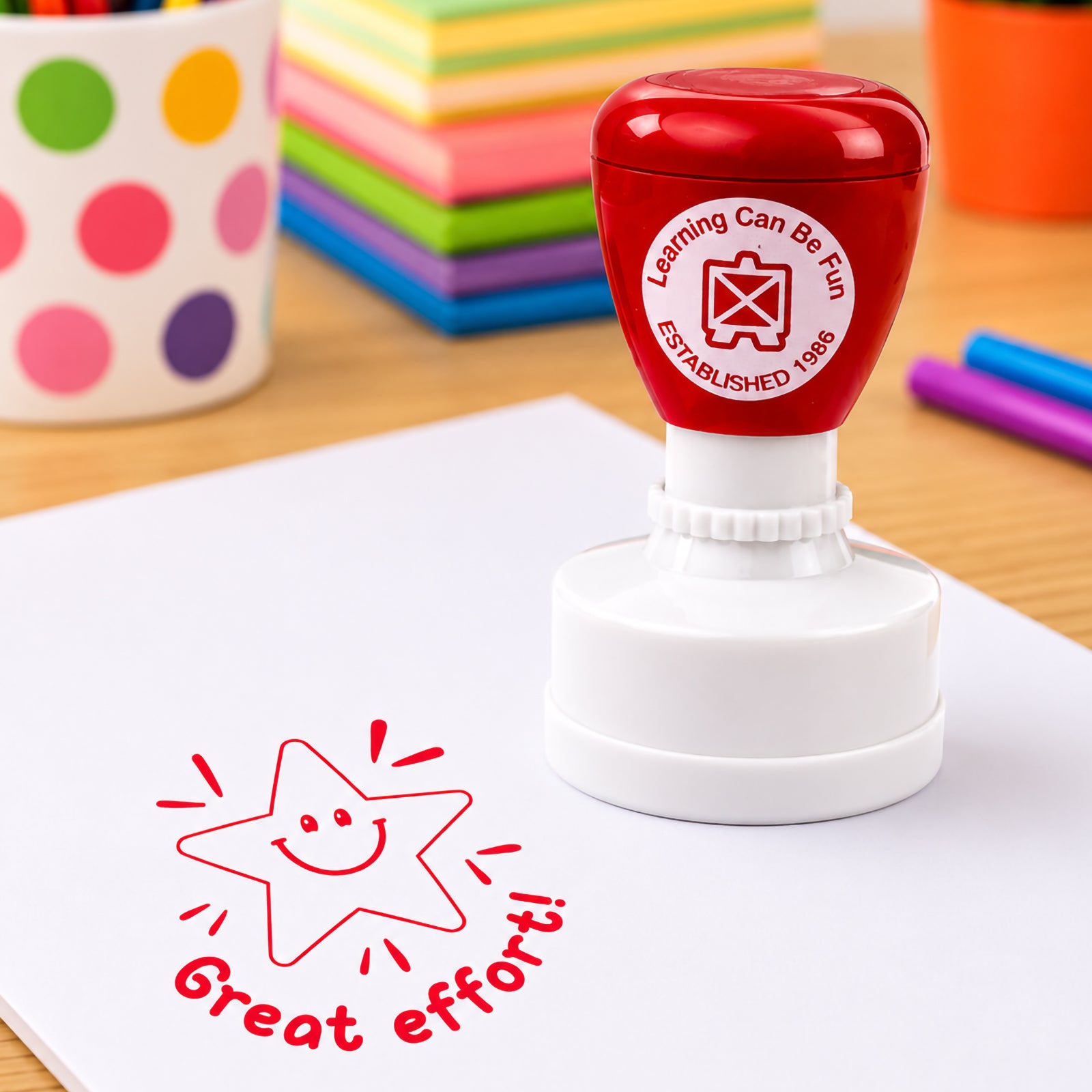Great Effort Merit Stamp shown in a colourful classroom environment with stacked notebooks, markers and educational supplies. The bold red stamp impression stands out clearly on student work.