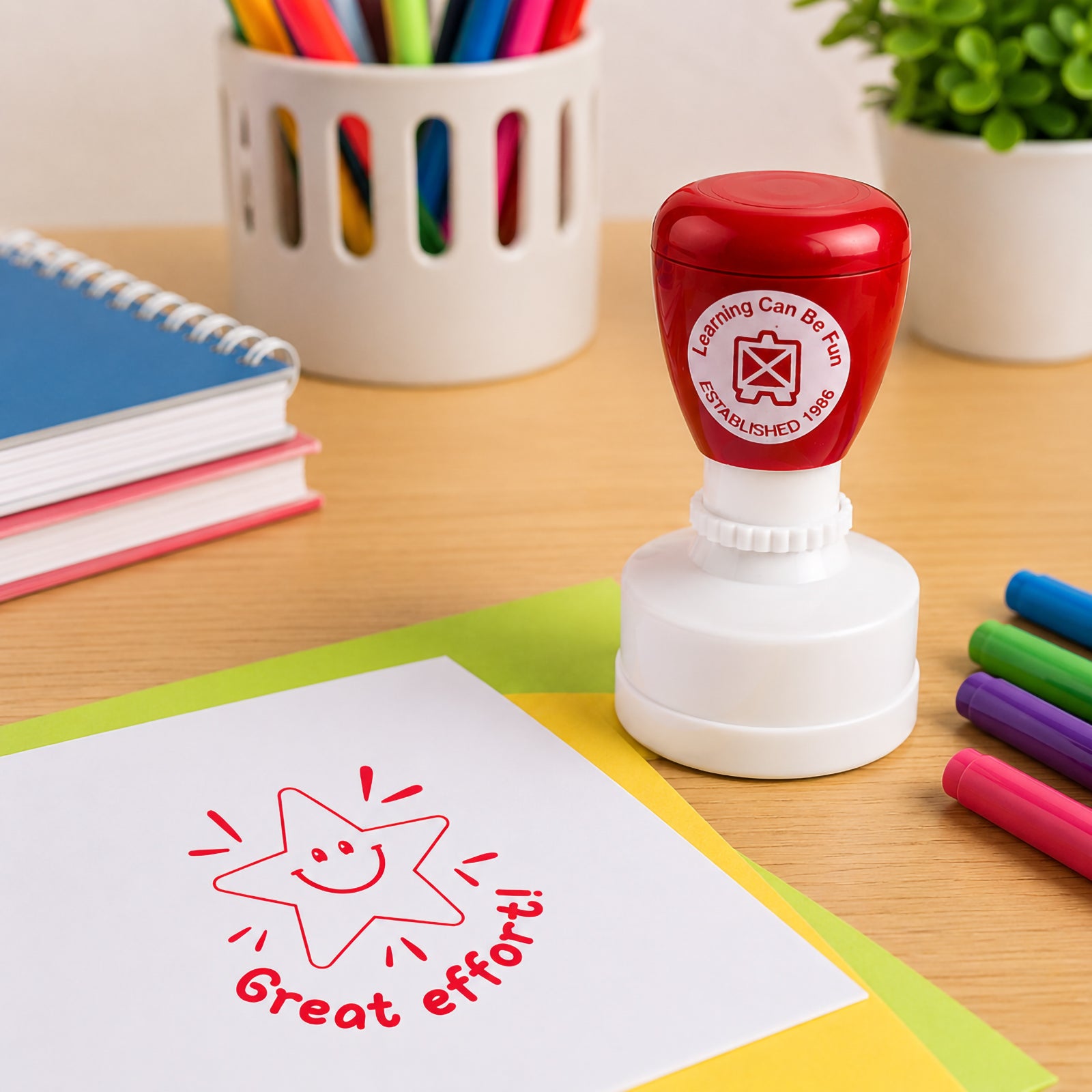 Self inking Great Effort reward stamp positioned on a desk surrounded by stationery and learning resources. The cheerful smiling star design creates a vibrant red impression ideal for motivating students.