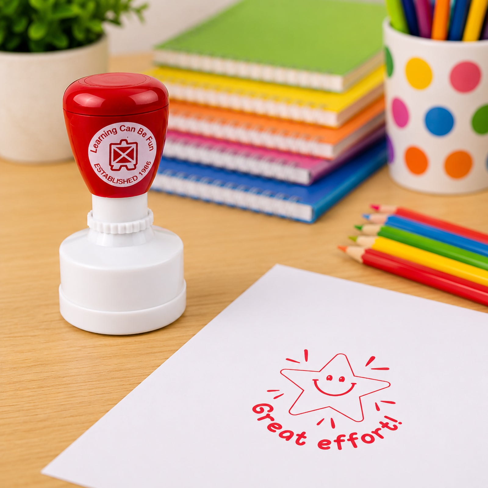 Learning Can Be Fun Great Effort Merit Stamp displayed beside colourful pencils and exercise books with a bright red Great Effort! star impression stamped onto white paper. The classroom themed setting highlights positive student recognition.