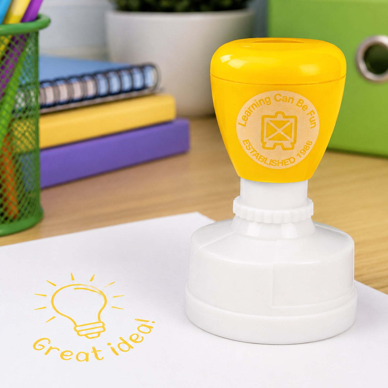 Great Idea Merit Stamp shown in a colourful educational setting with folders, notebooks and pencil holders. The vibrant yellow stamp impression stands out clearly on student work and classroom activities.