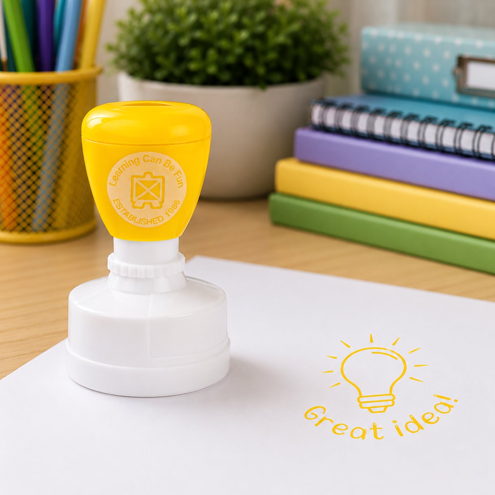 Learning Can Be Fun Great Idea Merit Stamp positioned on a classroom desk beside stationery and colourful notebooks. The yellow lightbulb design creates a clear Great Idea! impression on white paper, highlighting creativity and student achievement.