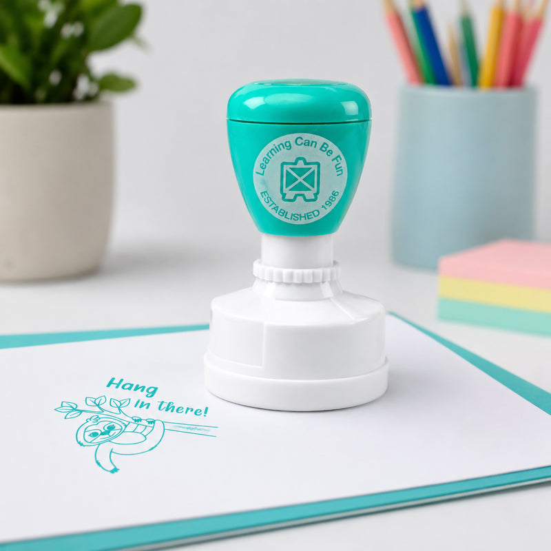 Close up view of the aqua merit stamp standing on a workbook beside the clear Hang in There! sloth impression. The encouraging message promotes resilience, confidence and positive learning habits.