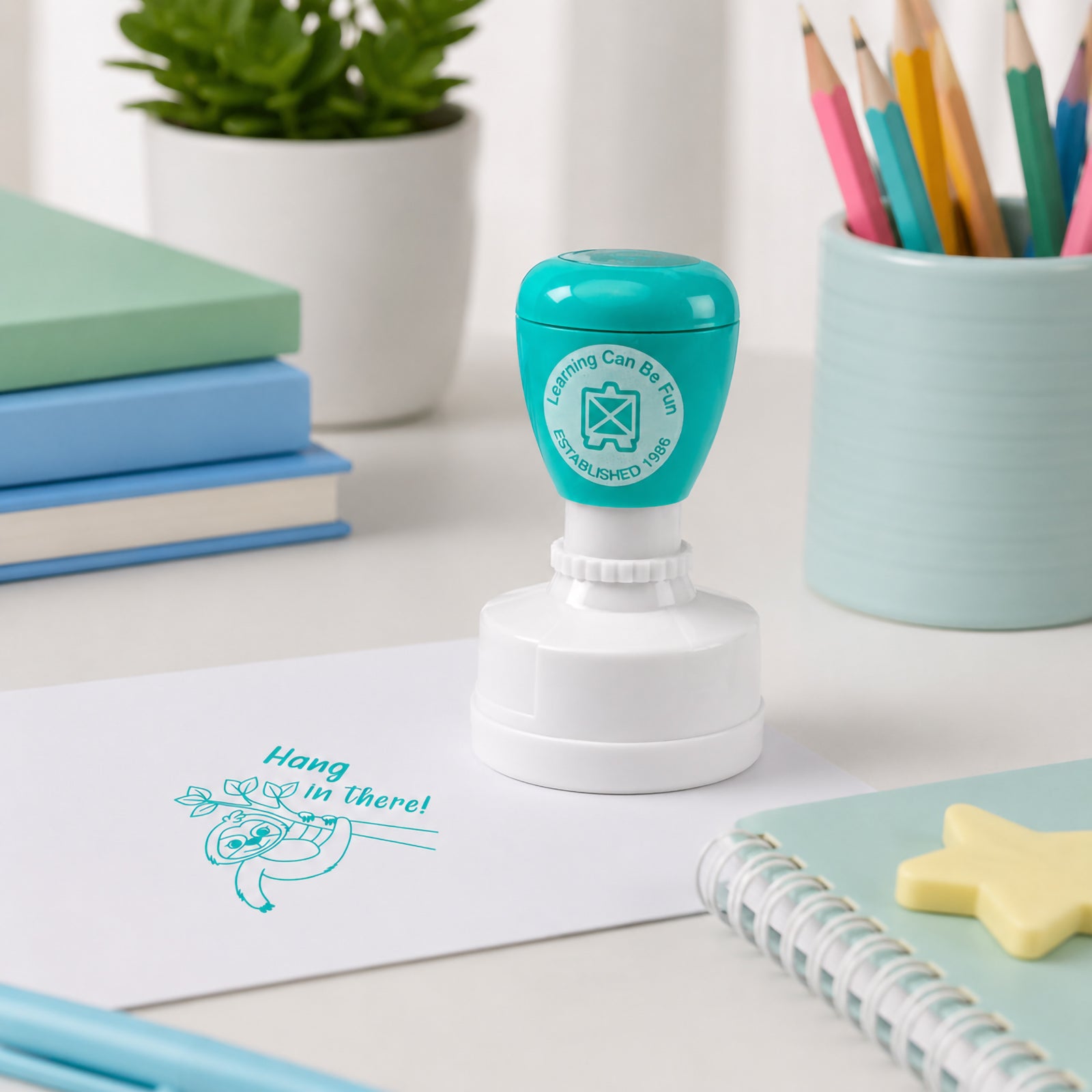 Self inking Hang in There reward stamp positioned on a classroom workspace with books, pencils and learning materials. The motivational sloth design helps teachers recognise perseverance and determination in a fun way.