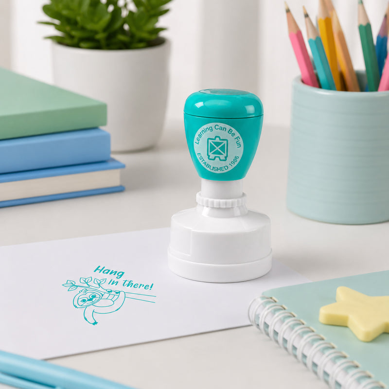 Self inking Hang in There reward stamp positioned on a classroom workspace with books, pencils and learning materials. The motivational sloth design helps teachers recognise perseverance and determination in a fun way.