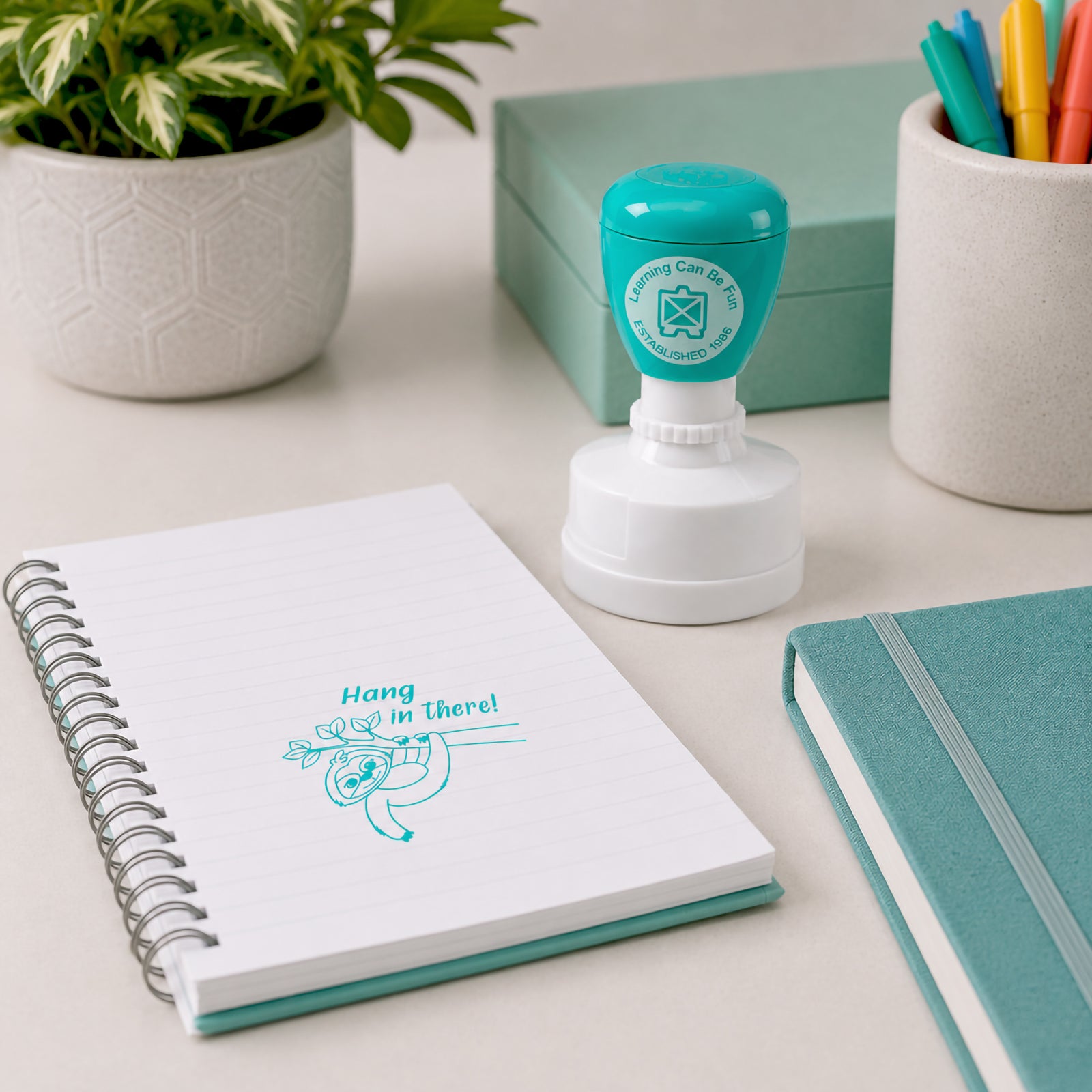 Learning Can Be Fun Hang in There Merit Stamp displayed beside notebooks and stationery on a modern desk. The cheerful aqua coloured sloth impression appears on a spiral notebook page, providing positive encouragement for students working through challenges.