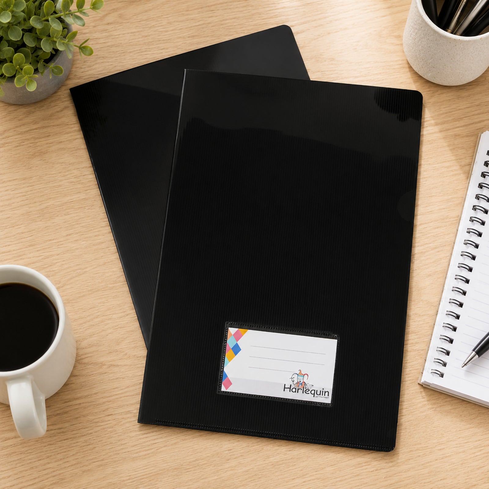 Top view of two Harlequin black letter files arranged on a wooden office desk beside a notebook, coffee cup, pen holder and decorative plant. The image showcases the smooth black finish and front card pocket for efficient document organisation.
