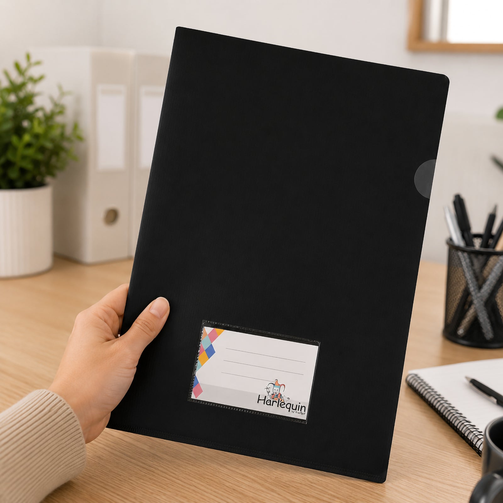 A person holds a Harlequin black letter file with a front card pocket in a modern office environment. The file is displayed above a timber desk with stationery, notebooks and desktop organisers, highlighting its professional appearance and convenient everyday handling.

Image 2
