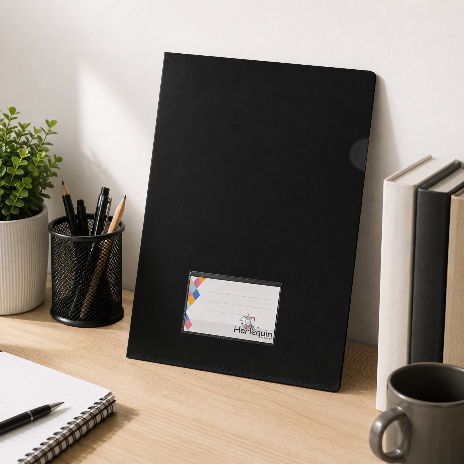 Harlequin black letter file standing upright against a wall on a clean office desk beside books, stationery and indoor plants. The professional workspace demonstrates how the file integrates neatly into everyday office filing and desktop organisation.
