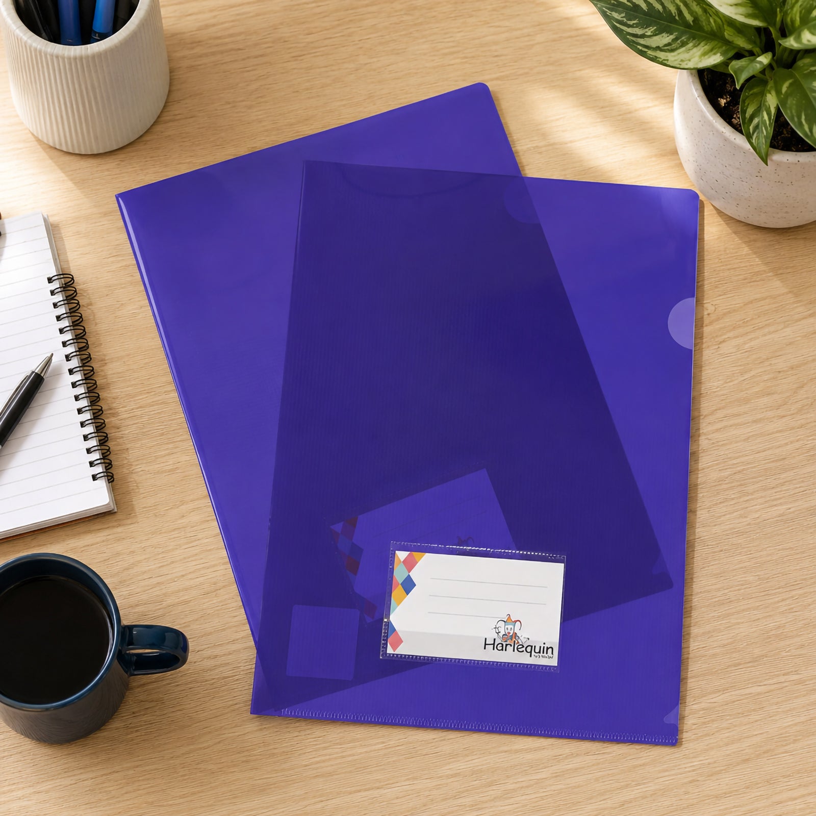 Top view of multiple Harlequin purple letter files arranged on a wooden office desk beside a notebook, coffee mug, pen holder and indoor plant. The image highlights the vibrant purple finish, front card pocket and practical organisation for busy workspaces.