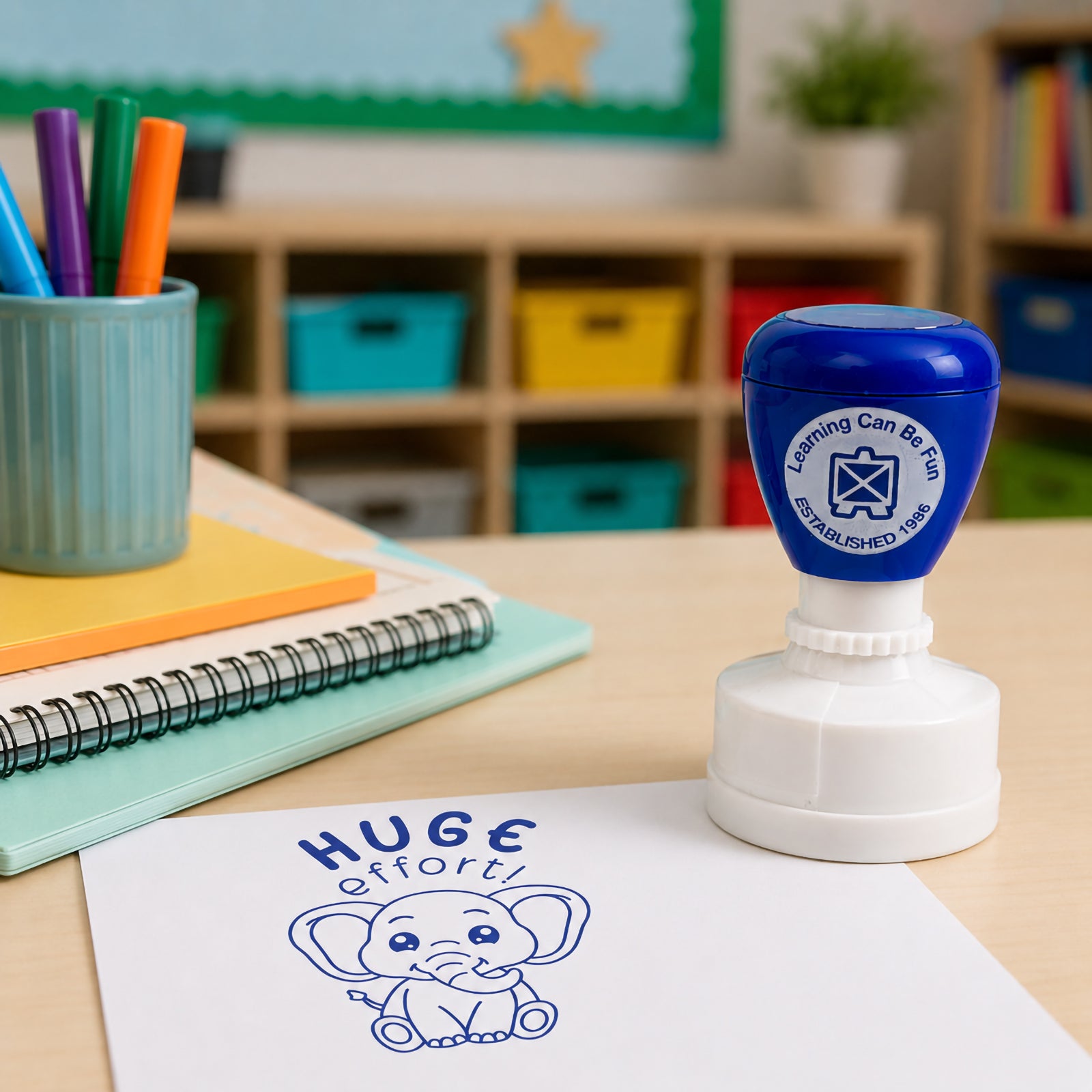Blue self inking Huge Effort merit stamp positioned on a classroom workspace with notebooks and stationery. The cheerful elephant design creates a positive and engaging reward for recognising perseverance and learning progress.