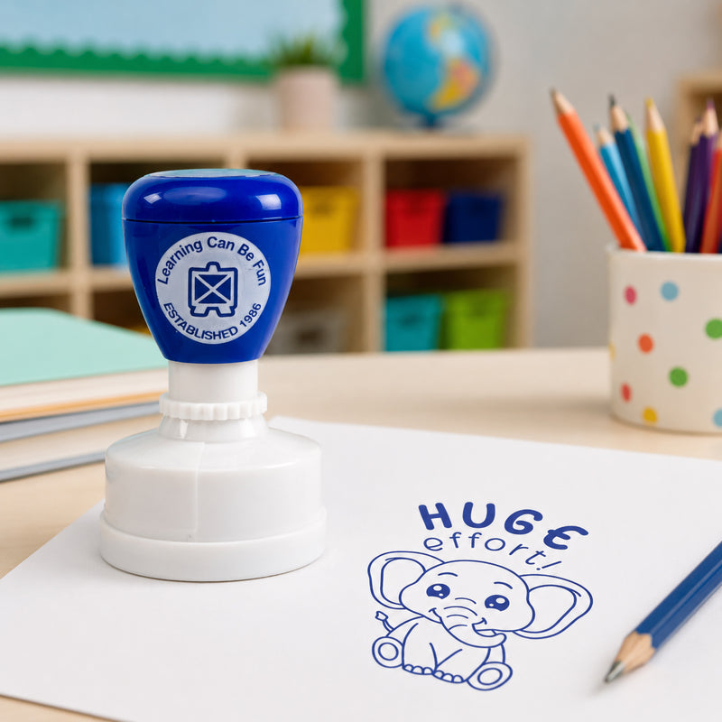 Learning Can Be Fun Huge Effort Merit Stamp displayed on a classroom desk beside books and coloured pencils. The blue elephant themed impression appears clearly on white paper, providing encouraging feedback for student achievement and effort.