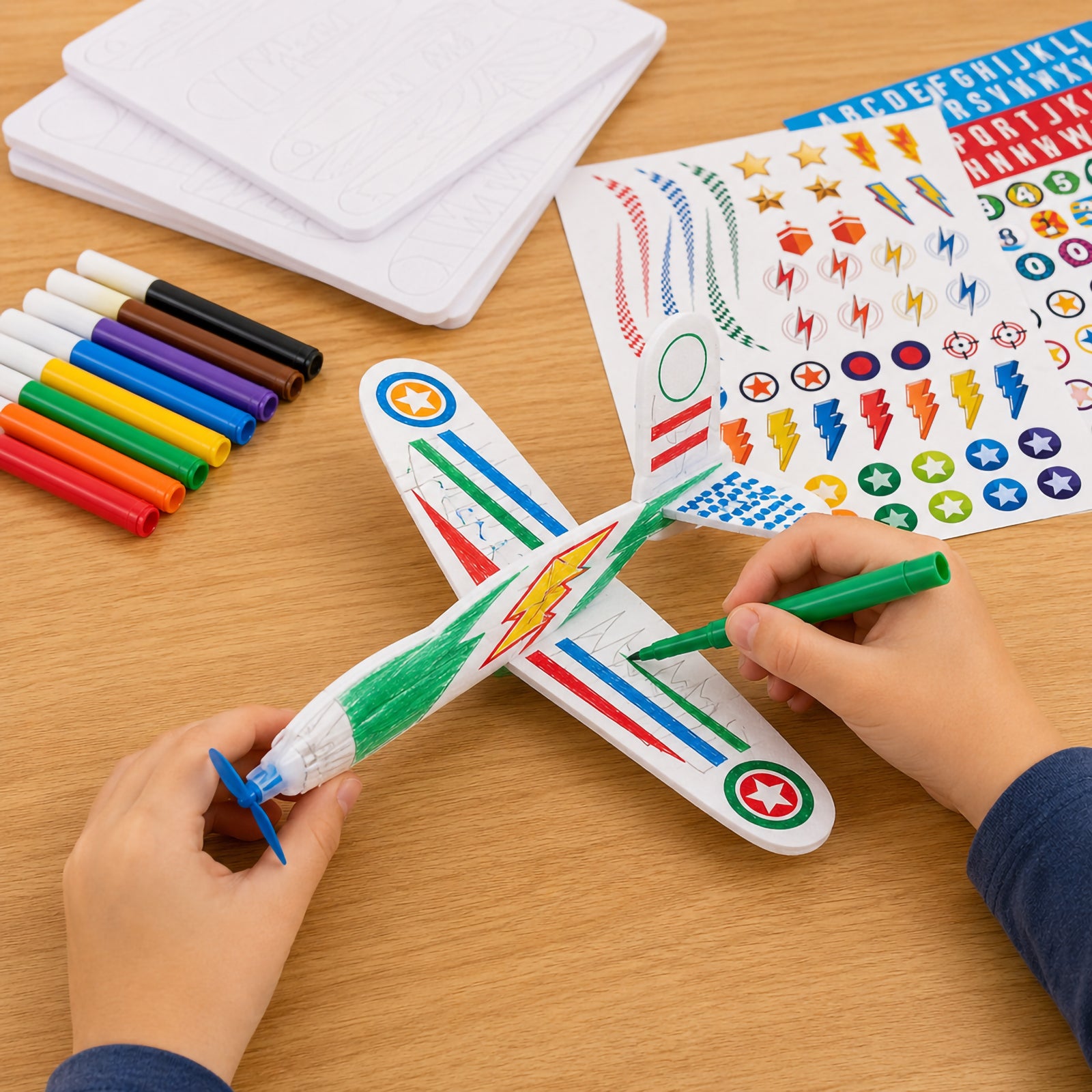 Close up of hands colouring a foam glider plane with a green marker while sticker sheets and coloured markers are displayed nearby. The image highlights the customisation process and creative freedom available when designing each aircraft.