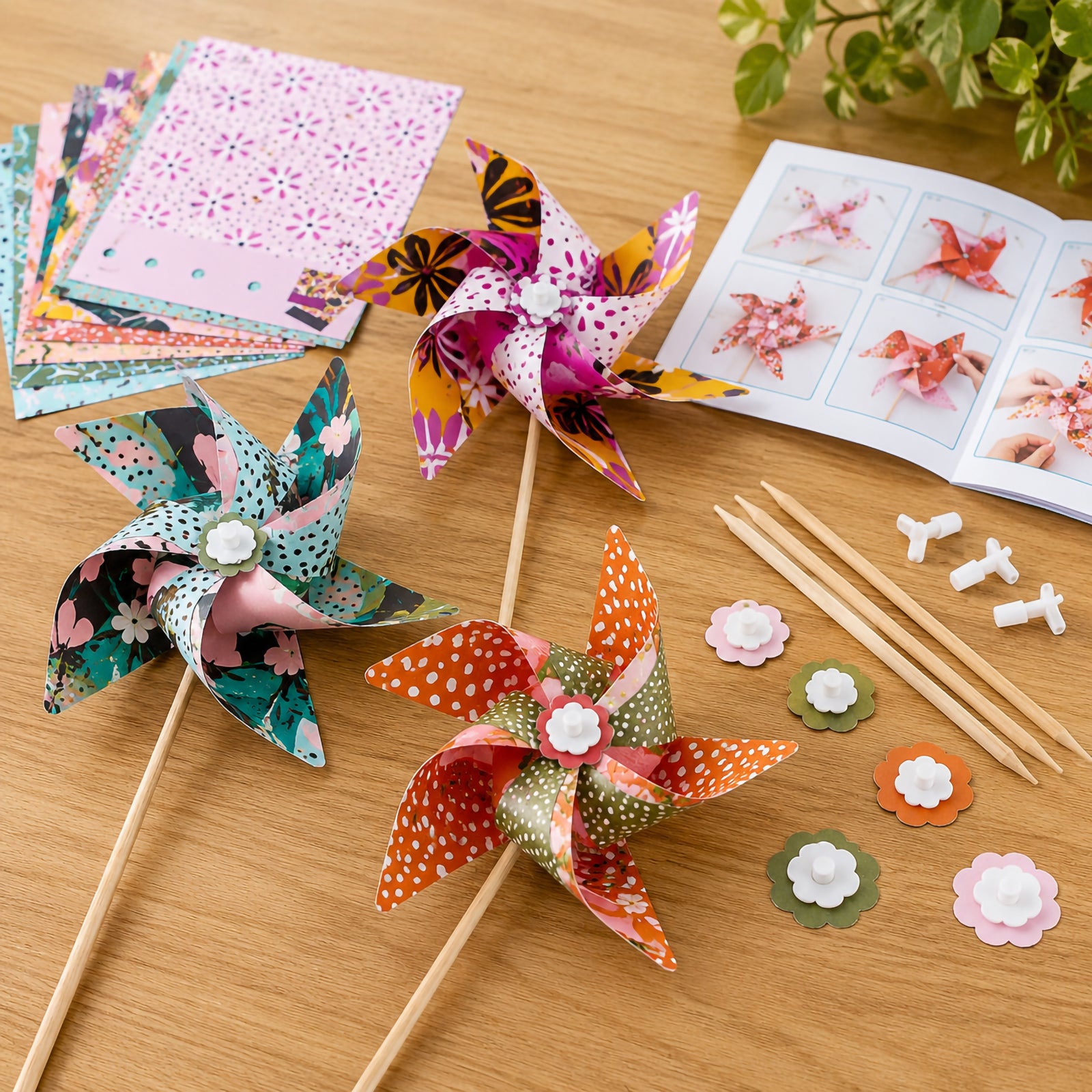 Three completed colourful windmills are displayed on a tabletop alongside printed paper templates, assembly pieces, wooden sticks and instruction booklet. The image highlights the creative crafting process and the vibrant finished windmill designs included in the kit.