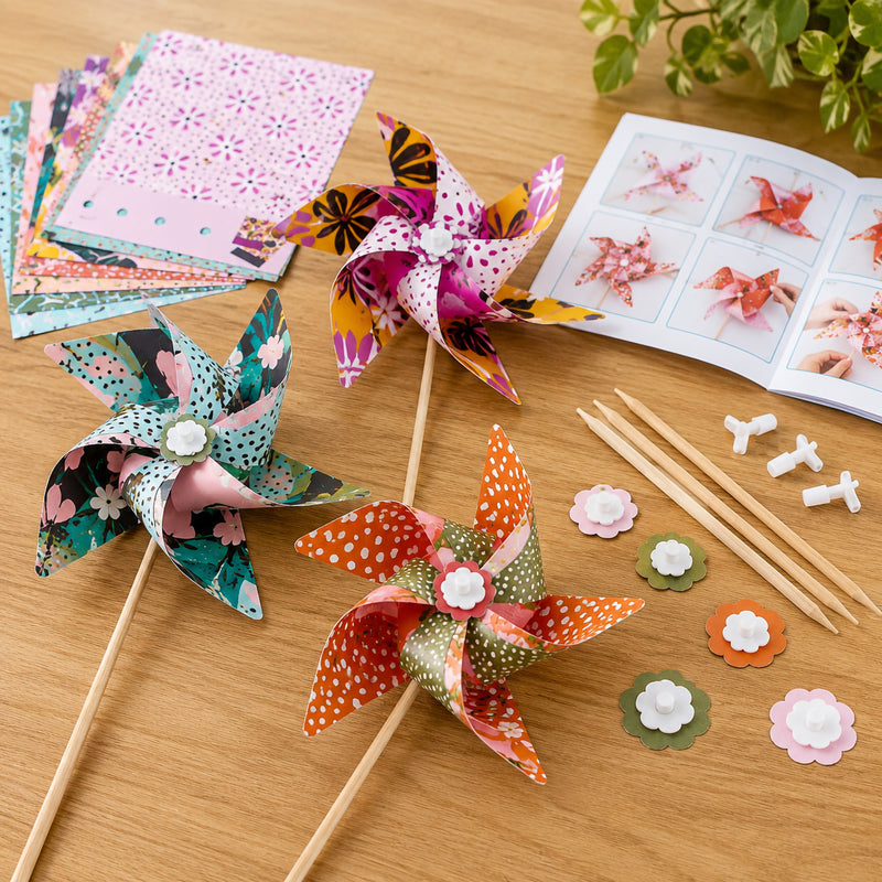 Three completed colourful windmills are displayed on a tabletop alongside printed paper templates, assembly pieces, wooden sticks and instruction booklet. The image highlights the creative crafting process and the vibrant finished windmill designs included in the kit.