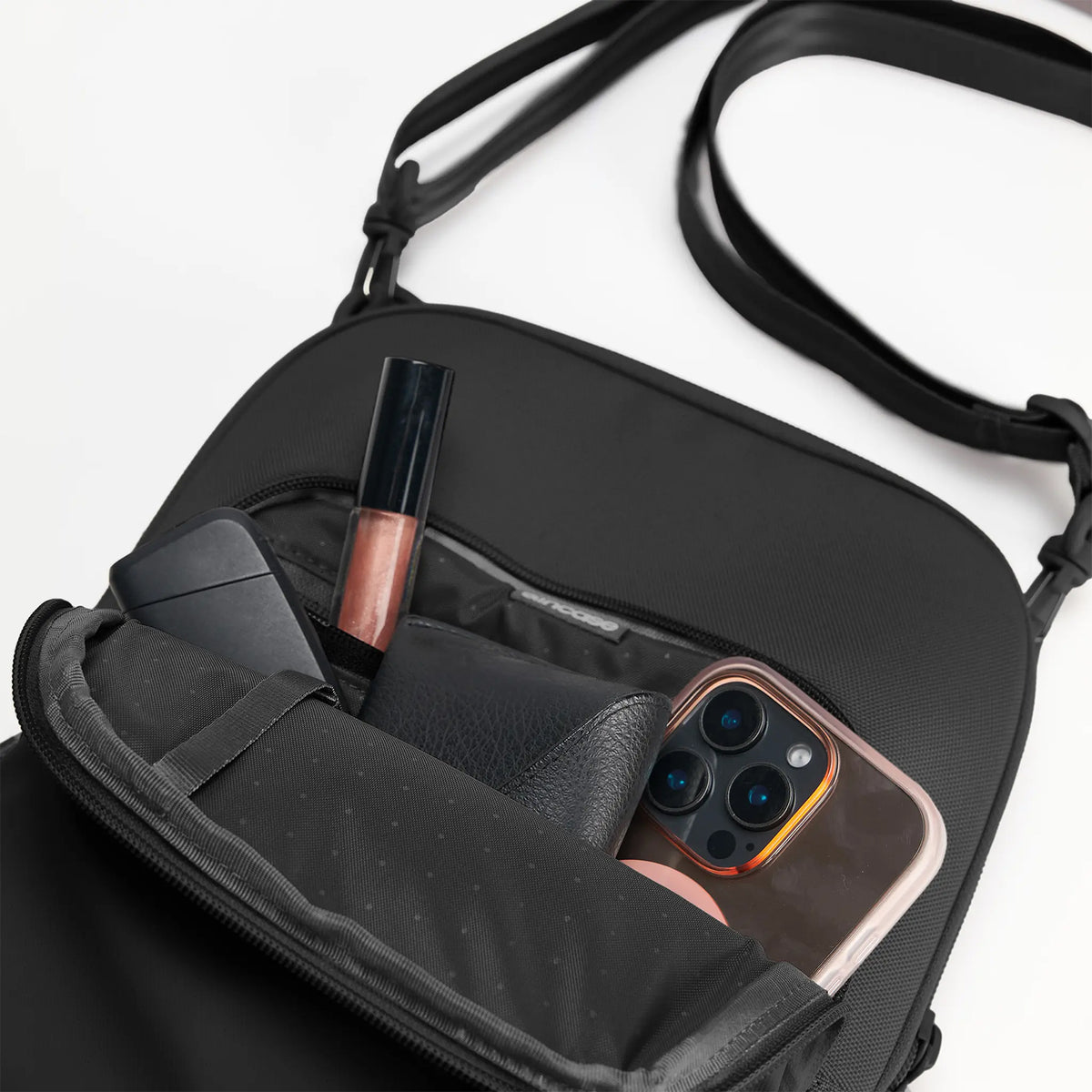 The Incase A.R.C. Crossbody Bag displayed with the front compartment open and filled with daily essentials including a smartphone, wallet, portable charger, cosmetics and accessories. Internal organiser pockets keep items neatly arranged while the compact design maximises usable storage for commuting, travel and everyday carry.