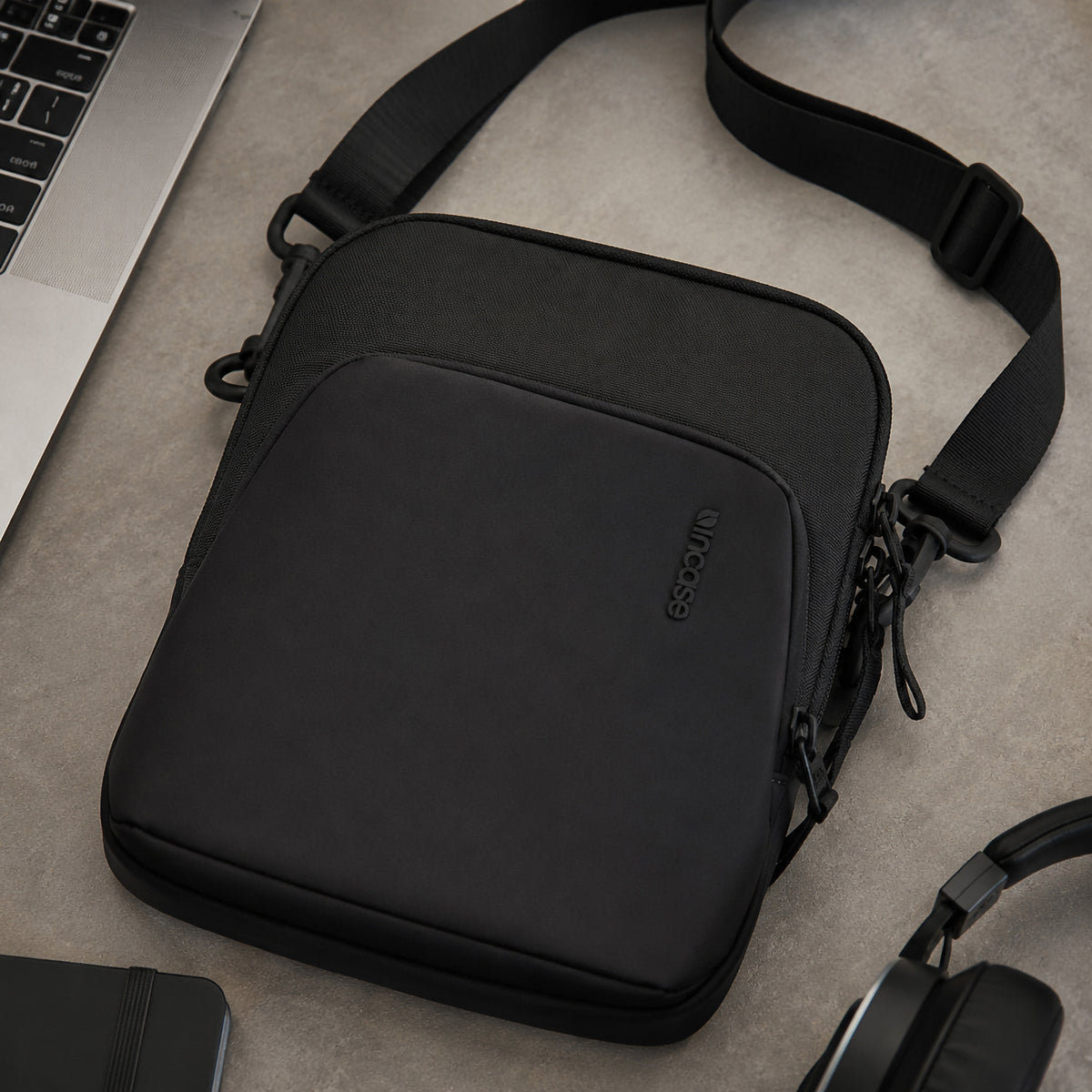Top down workspace view featuring the Incase A.R.C. Crossbody Bag beside a laptop, headphones and desk accessories. The image showcases the premium recycled polyester exterior, water resistant front panel and clean professional appearance, making it an ideal everyday carry bag for work or study.