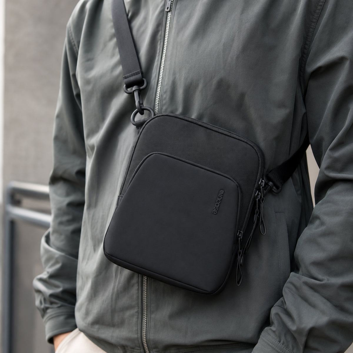 Lifestyle photograph of the Incase A.R.C. Crossbody Bag worn across the body with a modern grey jacket. The compact black crossbody sits comfortably against the chest, highlighting its adjustable shoulder strap, sleek minimalist styling and lightweight profile, making it ideal for commuting, travel and everyday urban use.