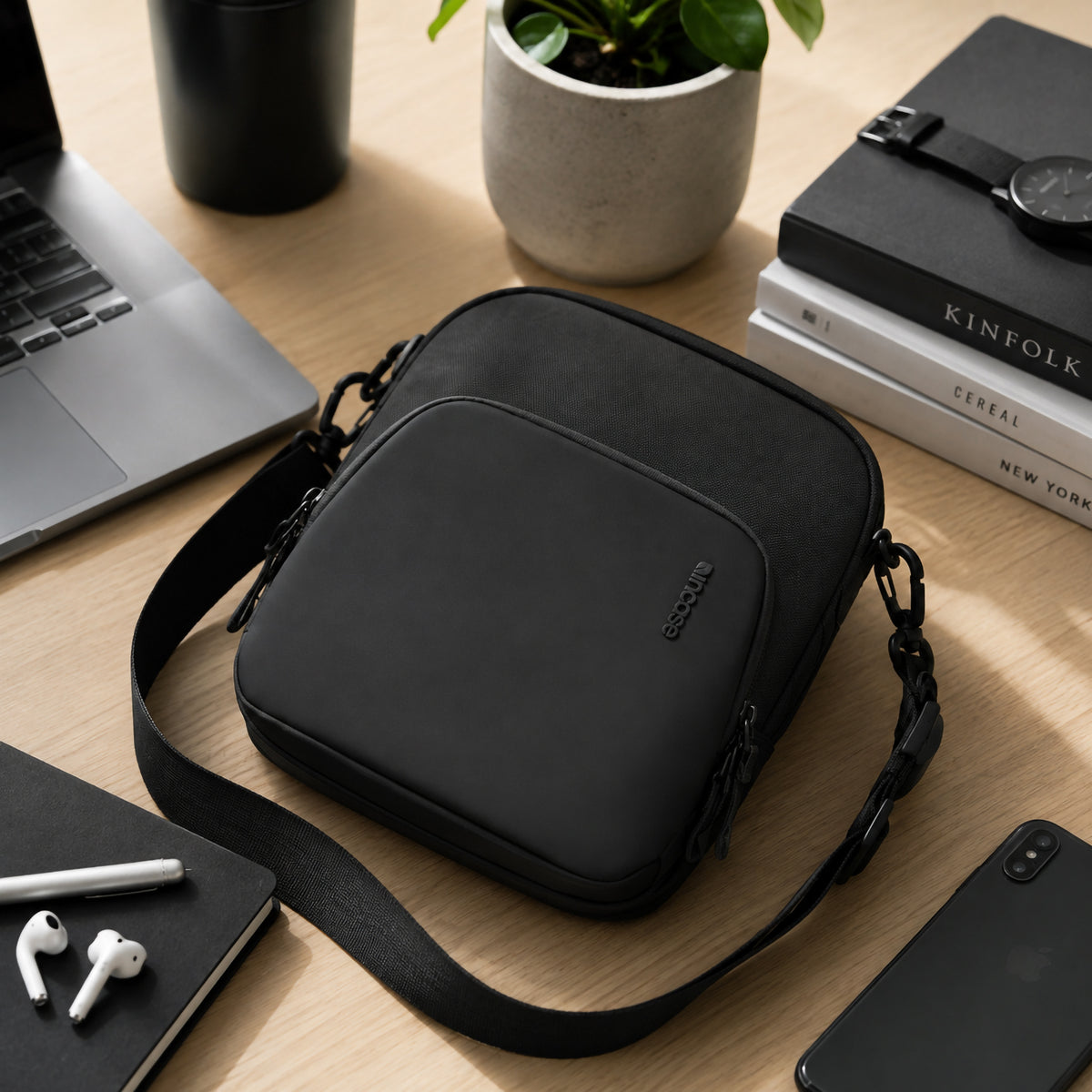 The Incase A.R.C. Crossbody Bag displayed on a tidy office desk surrounded by a laptop, notebook, smartphone, wireless earbuds, books and indoor plant. This lifestyle scene demonstrates how the bag integrates seamlessly into modern workspaces while keeping everyday essentials organised and within easy reach.