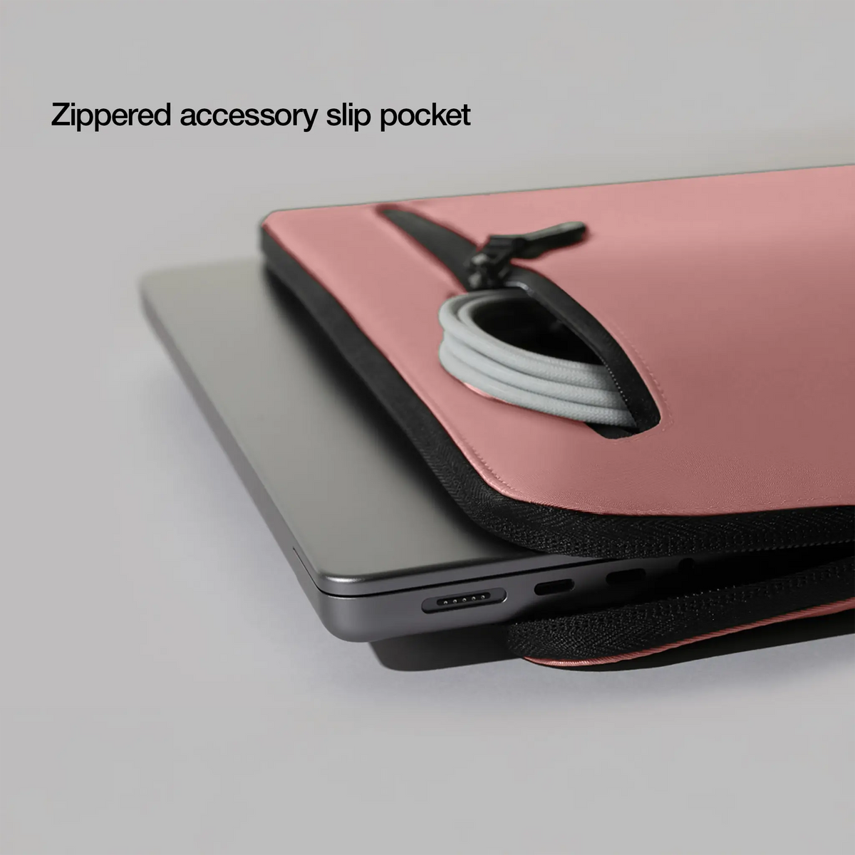Close-up of the Aged Pink Compact Sleeve front zip pocket holding a cable, with the caption Zippered accessory slip pocket