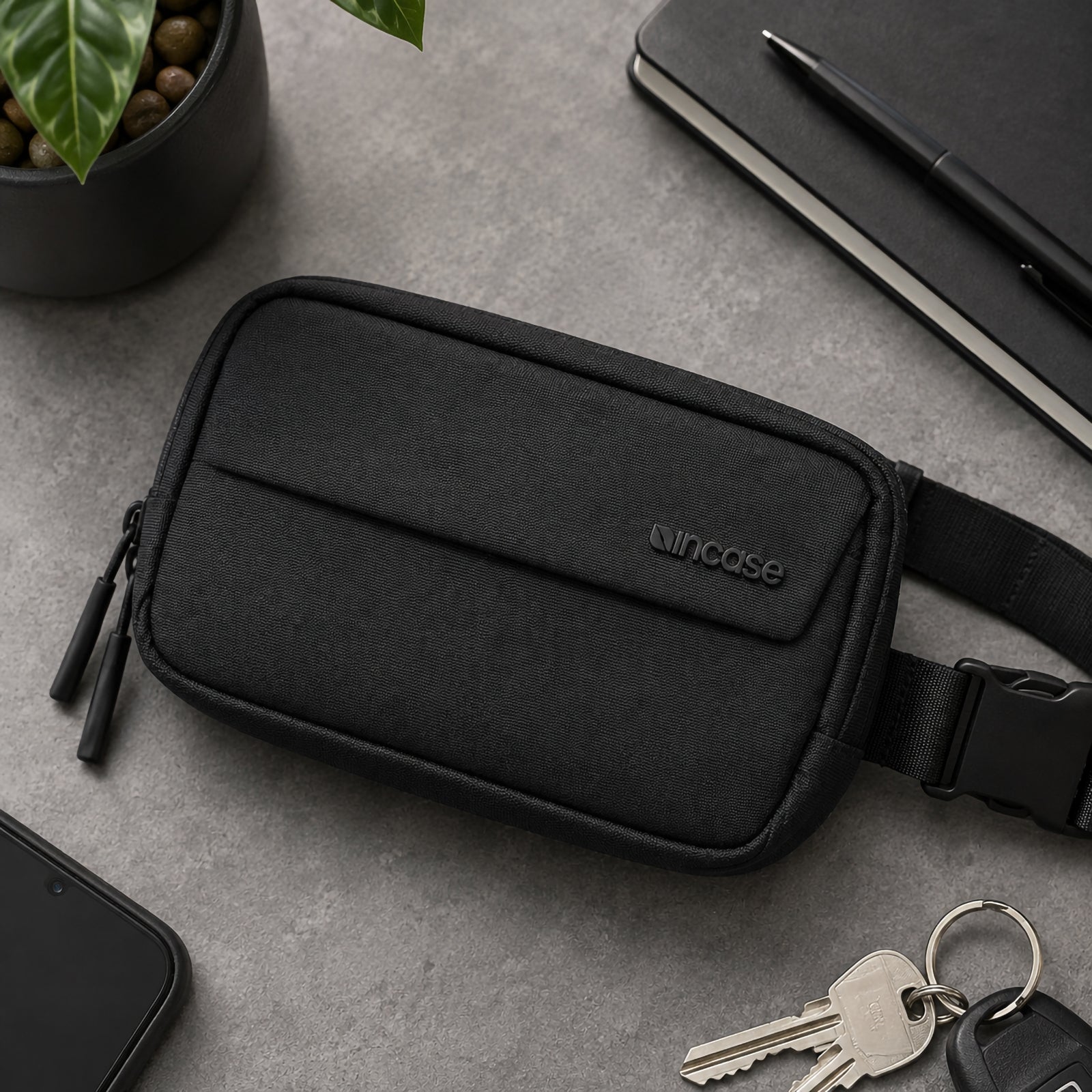 Top down desk flat lay of the Incase Crosstown Sling Black beside a smartphone, notebook, keys and indoor plant. The image highlights the premium textured exterior, clean design and compact footprint suitable for organised everyday carry.