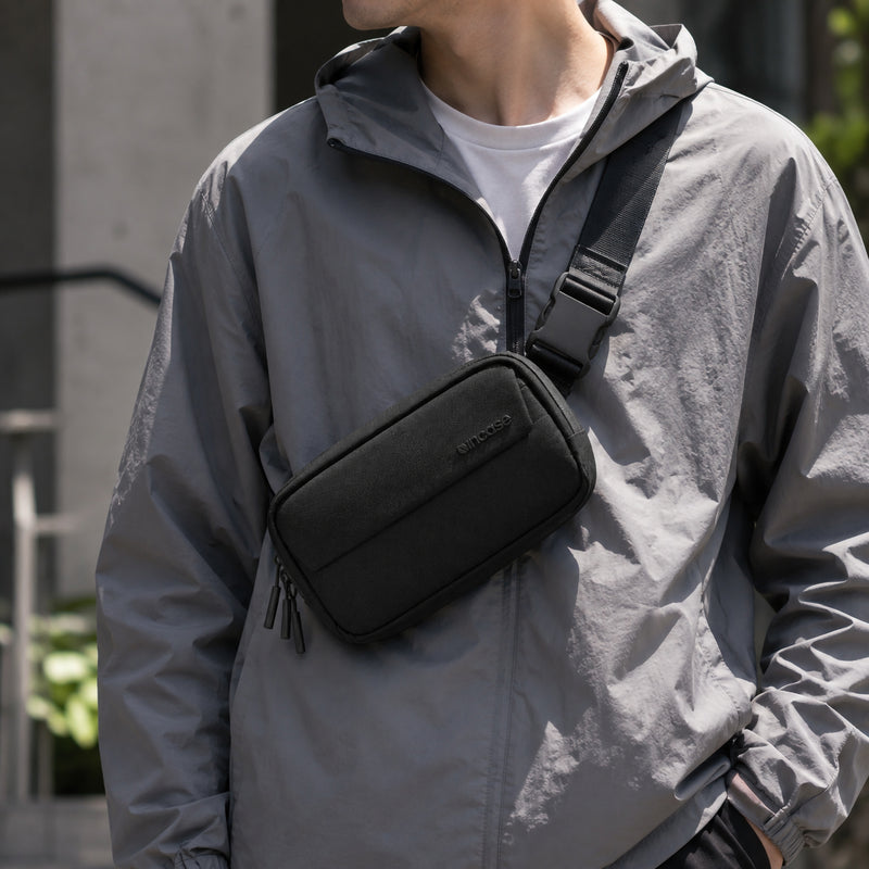 Lifestyle image showing the Incase Crosstown Sling Black worn across the chest with a lightweight grey jacket. The compact black sling sits securely against the body, demonstrating its minimalist profile and comfortable everyday crossbody fit for commuting and travel.